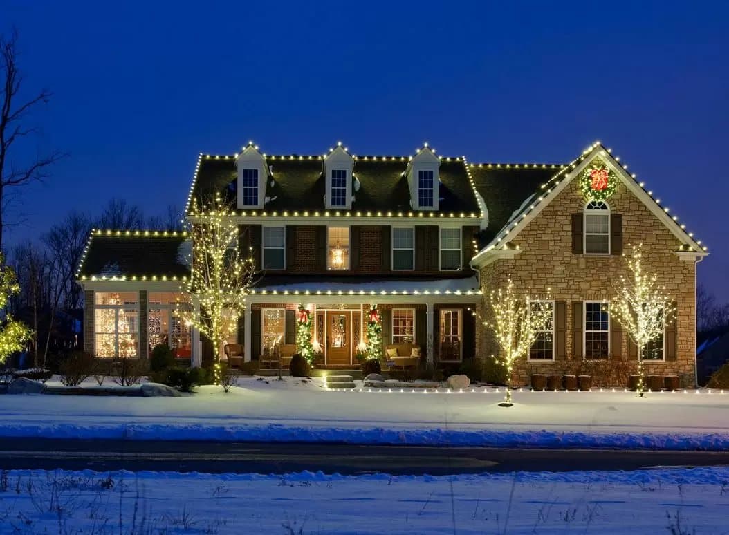 Colonial home with roofline lights and wrapped trees in snow